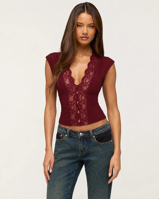 A woman modeling a sleeveless burgundy lace top with a deep V-neckline, paired with dark blue jeans, against a plain light gray background.