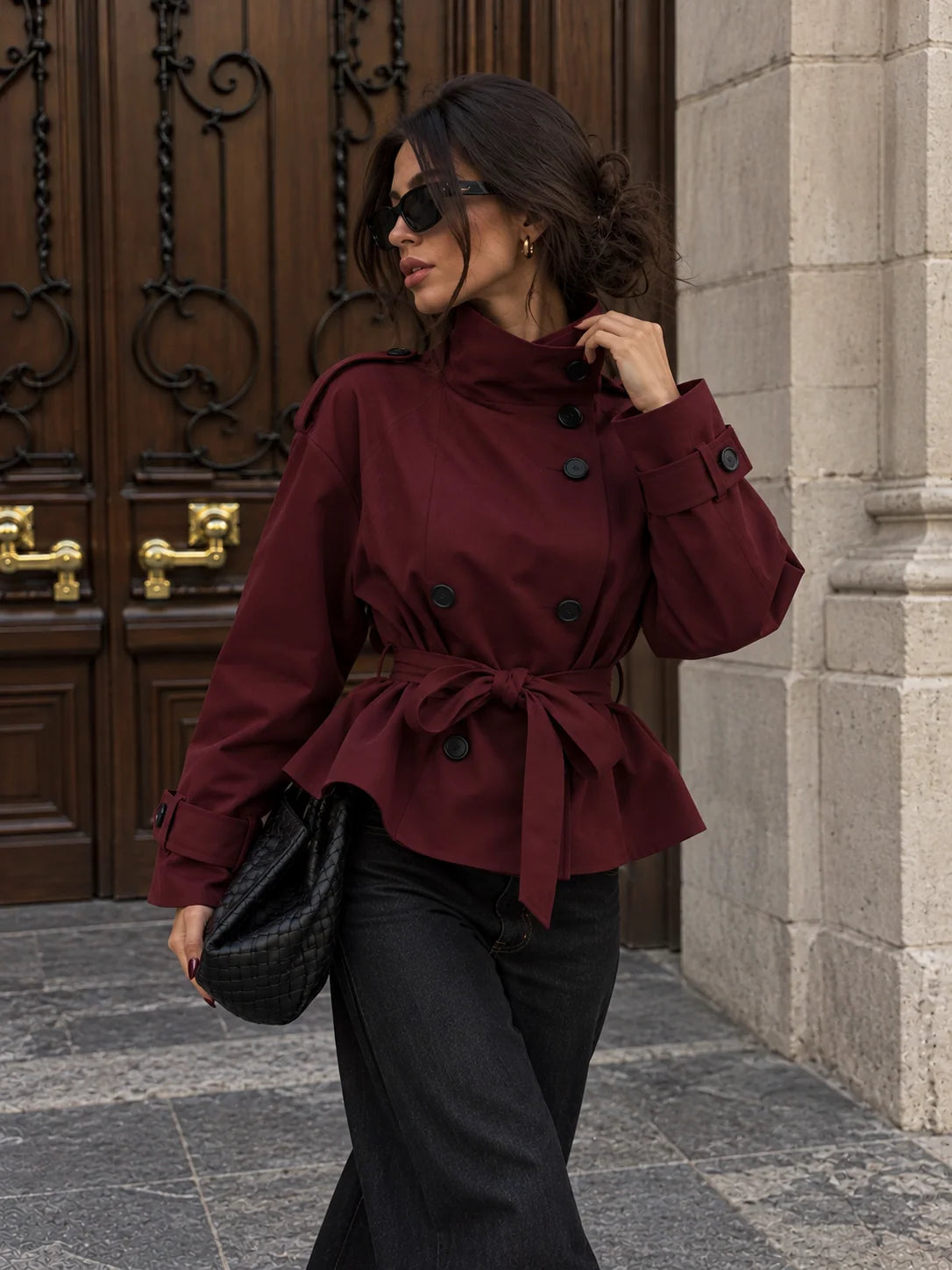 A woman wearing a burgundy belted double-breasted trench jacket and black pants, walking outdoors next to a stone wall and a dark wooden door.