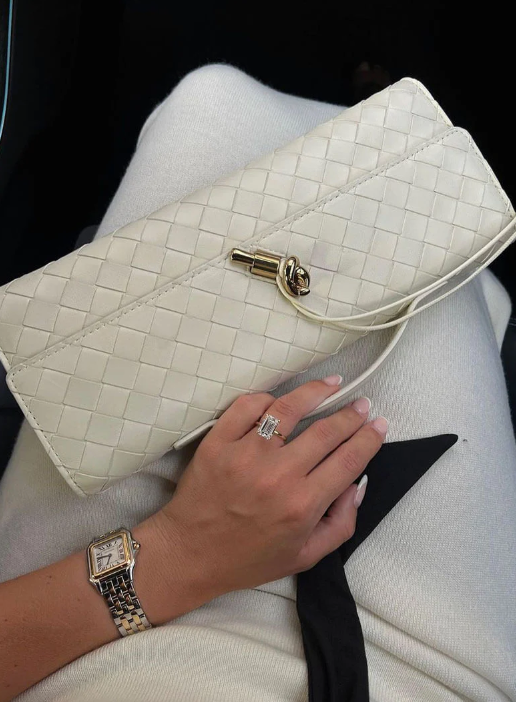 White woven handbag held by a hand with a ring, wearing a watch, on a light background.