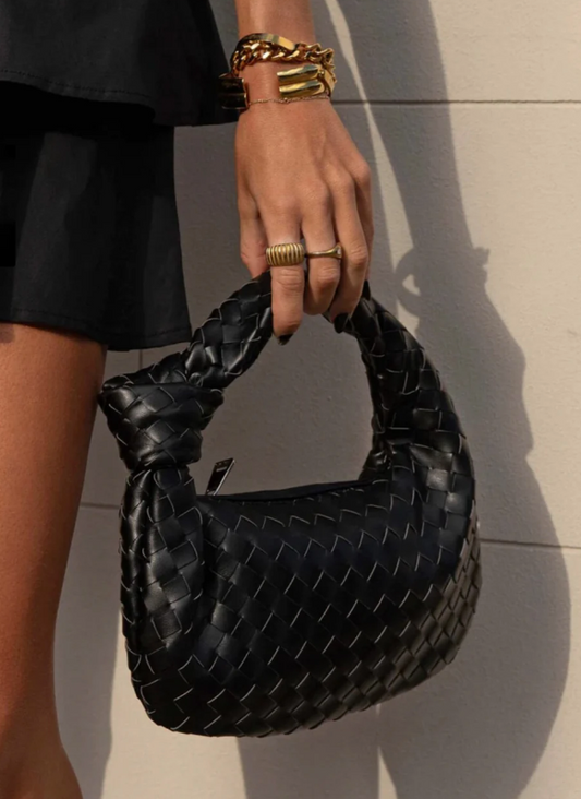Black woven handbag held by a person wearing gold bracelets and rings.