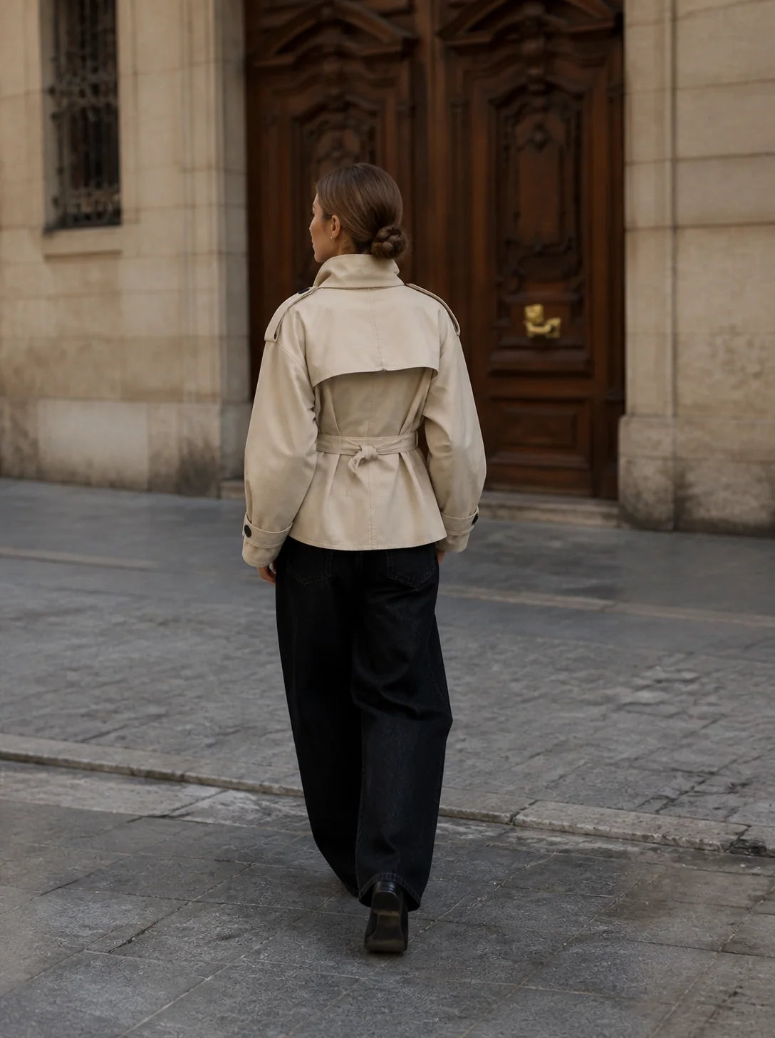 A woman viewed from behind walking on a stone street toward a large dark wooden door, wearing a light beige trench-style jacket with a belted waist and dark wide-leg pants.