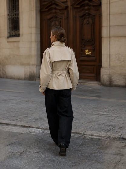 A woman viewed from behind walking on a stone street toward a large dark wooden door, wearing a light beige trench-style jacket with a belted waist and dark wide-leg pants.