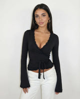 A woman modeling a black long-sleeve wrap top with a scalloped V-neckline and side tie, paired with white pants, against a plain white background.