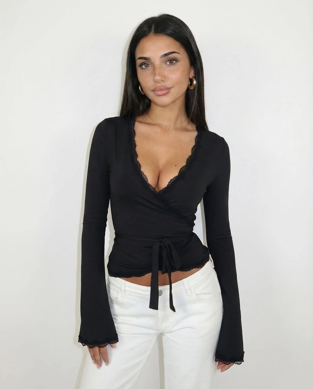 A woman modeling a black long-sleeve wrap top with a scalloped V-neckline and side tie, paired with white pants, against a plain white background.