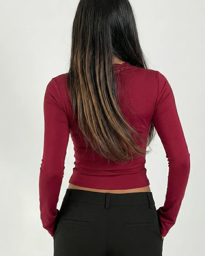A woman viewed from behind wearing a fitted long-sleeve crop top in deep burgundy and black pants, against a plain light gray studio background.