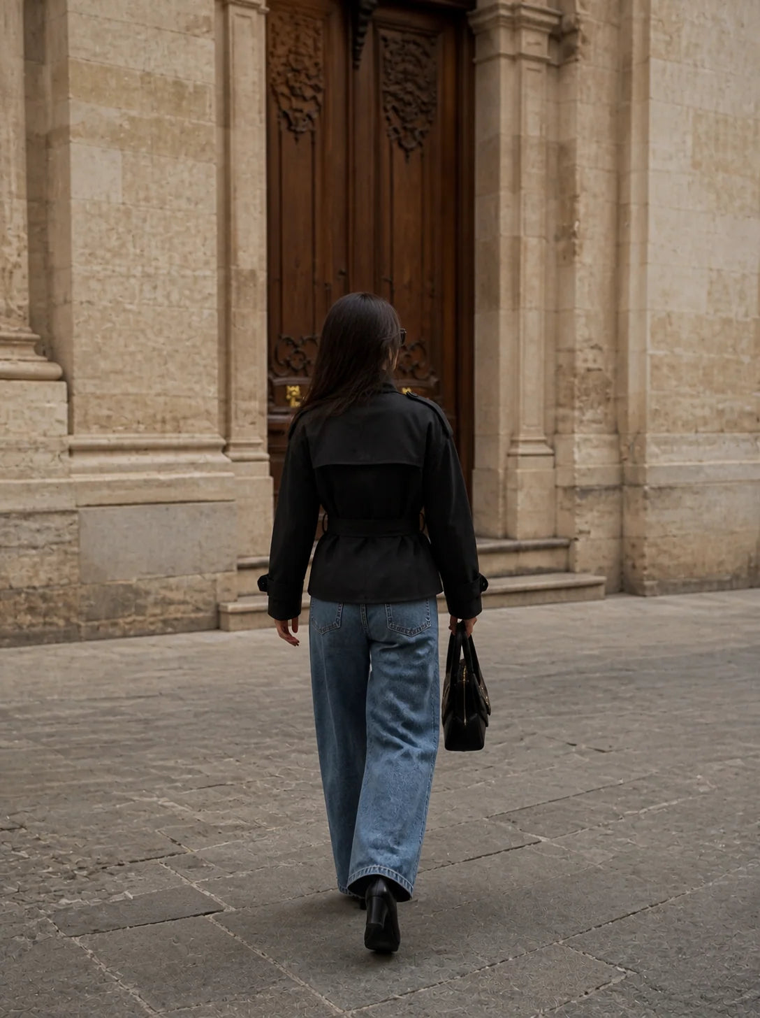 A woman viewed from behind walking toward a large wooden door in a stone building, wearing a black long-sleeve jacket, wide-leg light blue jeans, black boots, and carrying a black handbag.