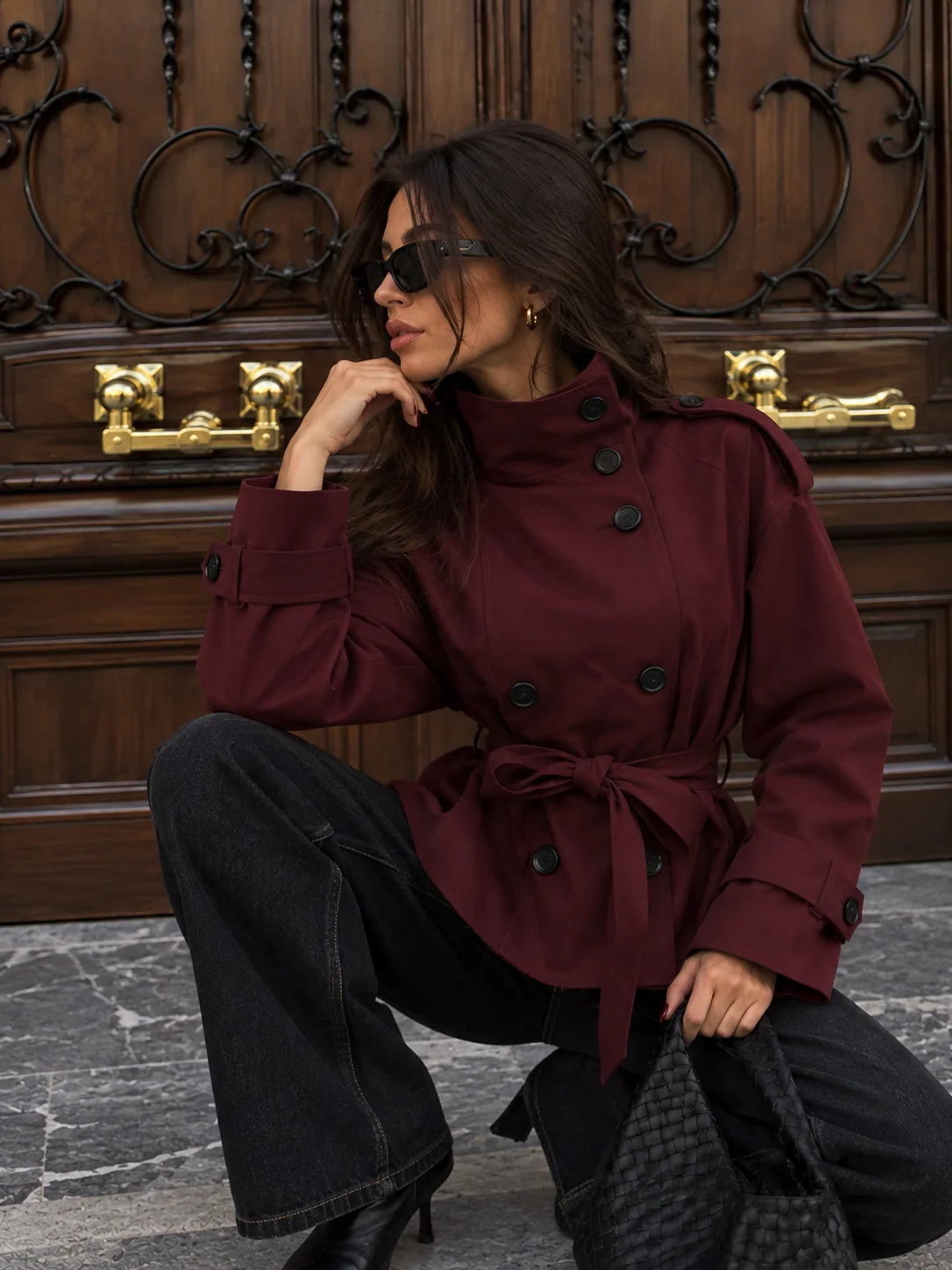 A woman wearing a burgundy belted double-breasted jacket, black wide-leg jeans, and sunglasses crouches in front of a dark wooden door with gold handles.