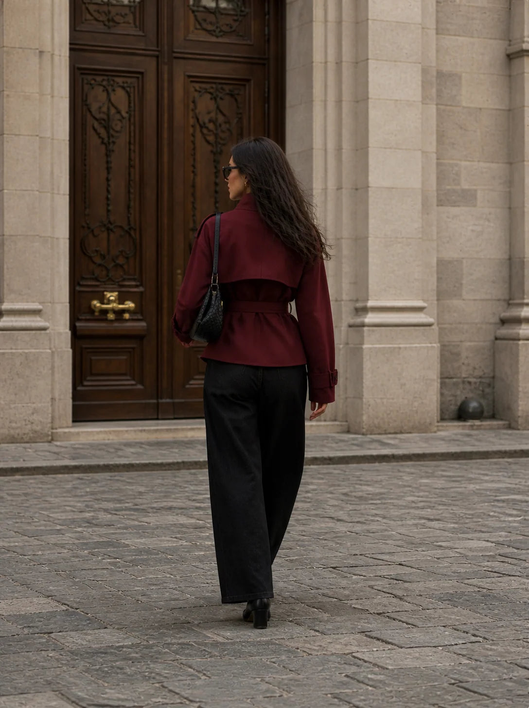 A woman viewed from behind walking toward a large ornate brown wooden door on a stone-paved street. She is wearing a dark burgundy cropped jacket with long sleeves, black wide-leg trousers, and carrying a black shoulder bag.