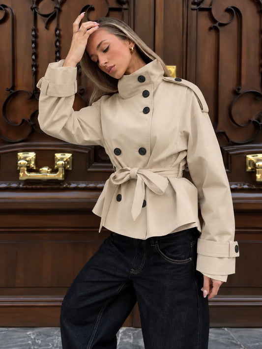 A woman wearing a beige double-breasted trench coat with a waist tie and dark wide-leg jeans, posing in front of a large dark wood door with gold handles.