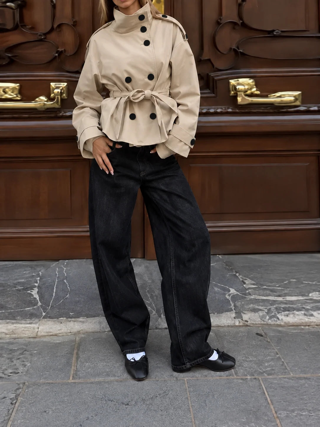 A model wearing a beige double-breasted trench jacket with a tie waist, paired with wide-leg black jeans and black loafers, standing in front of a dark brown wooden door.