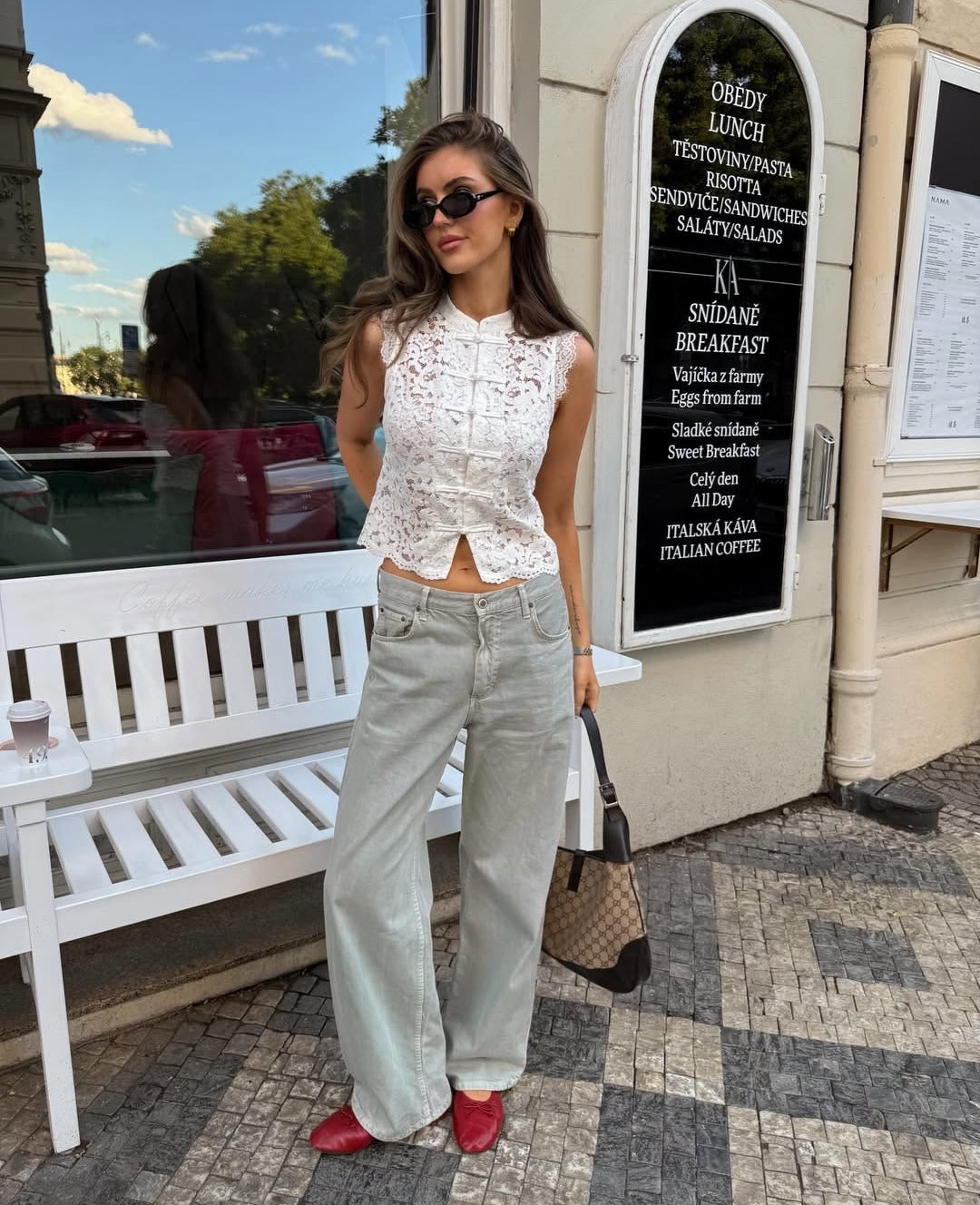 A woman stands outdoors on a sidewalk in front of a cafe storefront, wearing a sleeveless white lace top, light gray wide-leg jeans, red pointed shoes, and carrying a brown patterned handbag.