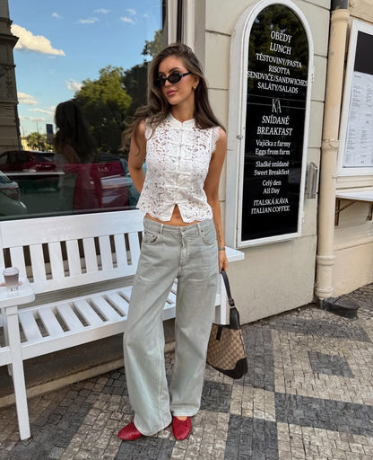 A woman stands outdoors on a sidewalk in front of a cafe storefront, wearing a sleeveless white lace top, light gray wide-leg jeans, red pointed shoes, and carrying a brown patterned handbag.