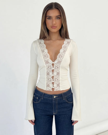 A woman modeling a fitted long-sleeve cream top with a lace front panel and deep V-neckline, paired with dark blue jeans, against a plain white studio background.
