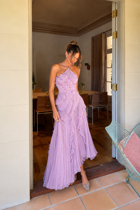 A woman wearing a lavender ruffled maxi dress stands in a doorway, holding out one side of the pleated skirt to show its flow. The background shows a warm-toned indoor room with wooden floors and furniture.