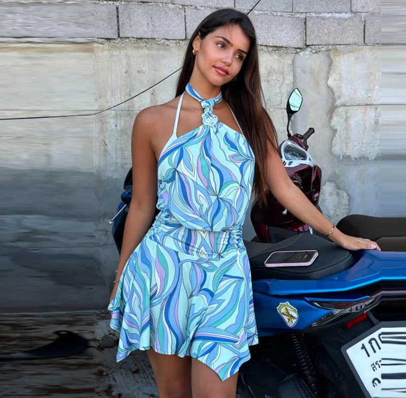 Woman in a blue patterned dress standing next to a motorcycle against a stone wall.