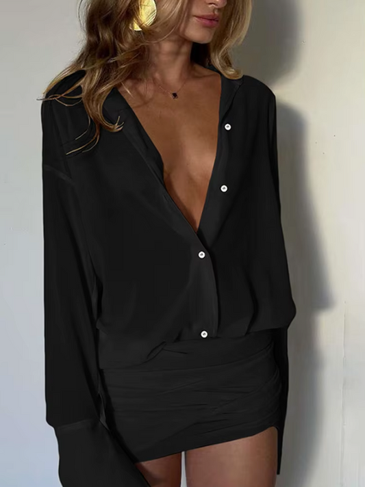 Woman wearing a black button-up shirt over a black skirt against a white wall.