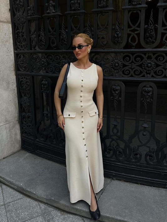 Woman in a long beige dress standing in front of an ornate black metal gate.